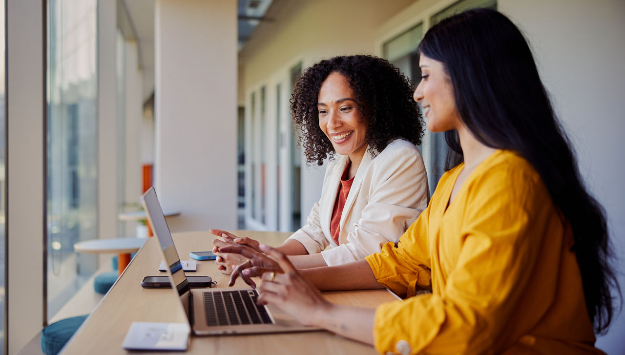 Two young businesswoman talking and smiling while working together on a laptop at a table in an office lounge