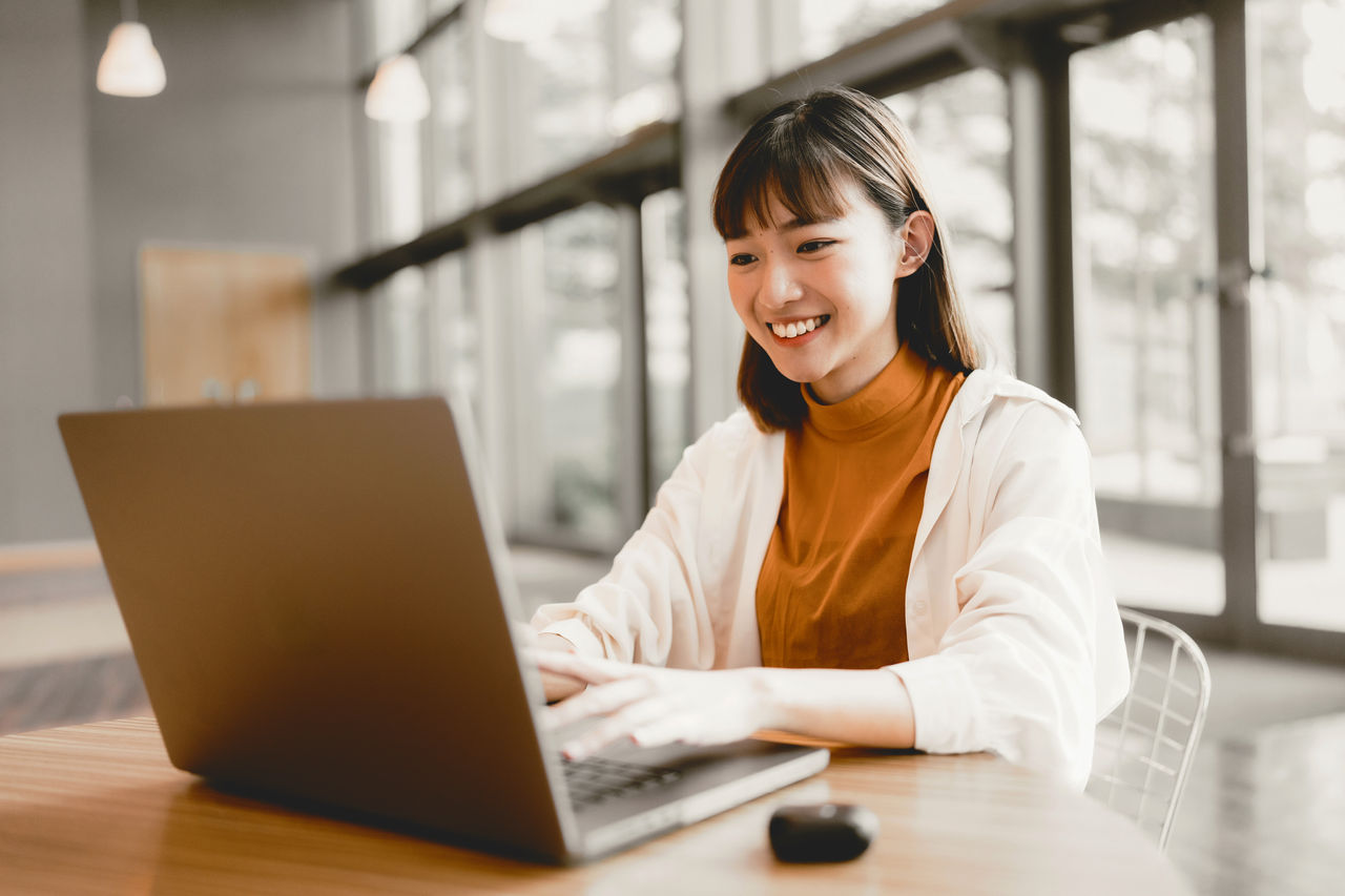 A medium shot of a young East Asian female adult working on her laptop while smiling. Both her hands are on the keyboard and she is using earphones. She is in the office with large windows on the back, allowing the sunshine to pass through.