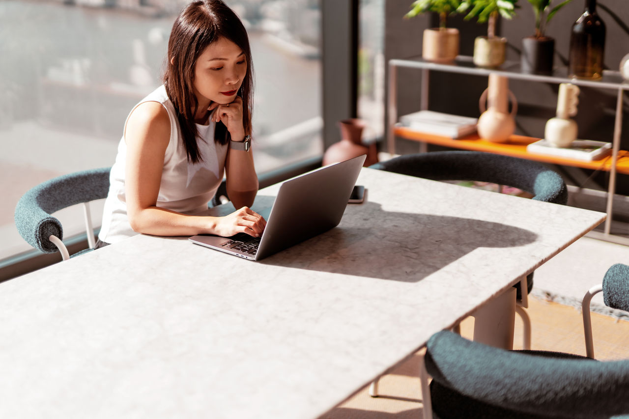 Young Asian woman working with laptop while sitting by the window with city view and sunlight.