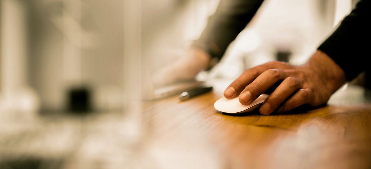 Closeup of a man's hand on a white computer mouse, placed on a wooden desk in an office setting. Blurred background, workplace concept
