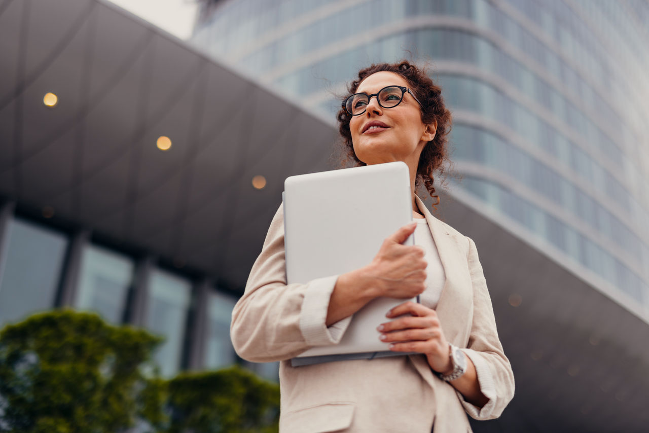 Smiling businesswoman in her 40s is holding a laptop in her arms while looking away and thinking about future projects in front of an office building