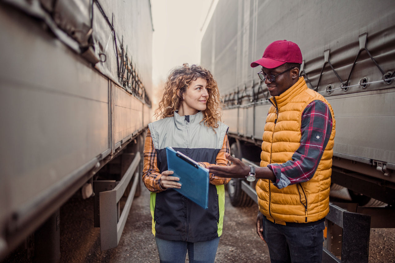 A female supervisor checks the truck and the truck driver before the trip begins. They are standing in the parking lot between two trucks