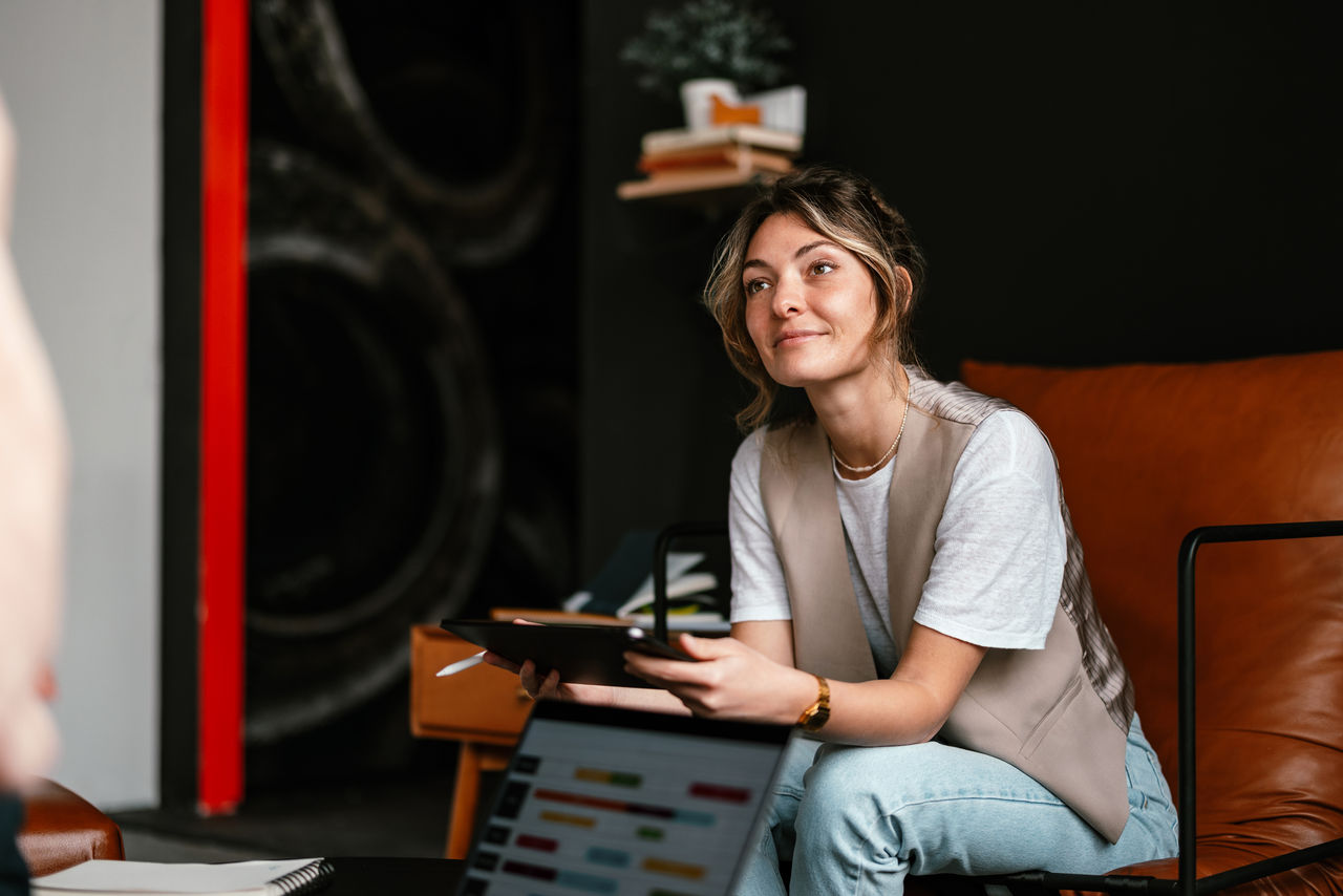 Close up shot of a beautiful woman sitting in a leather chair holding a digital tablet. She is listening to an anonymous person, looking away and smiling.