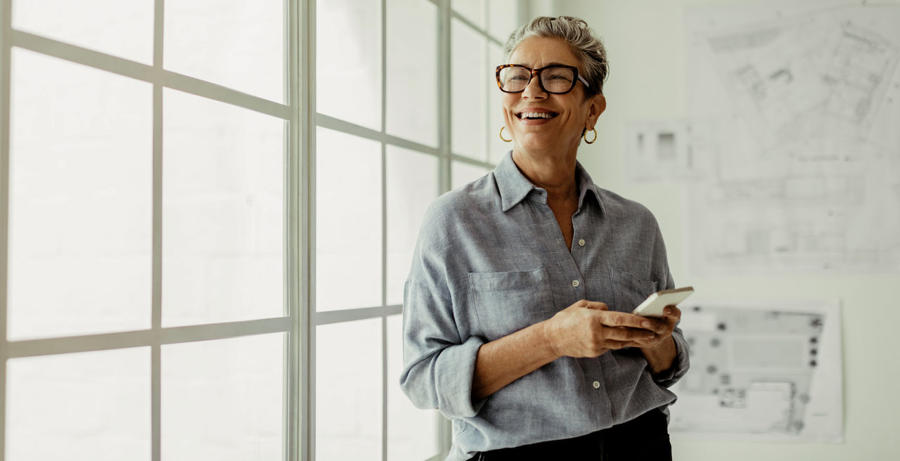 Mature business woman stands in an office, holding a smartphone and looking outside the window with a smile. Female designer using mobile technology to stay connected with her colleagues and clients.
