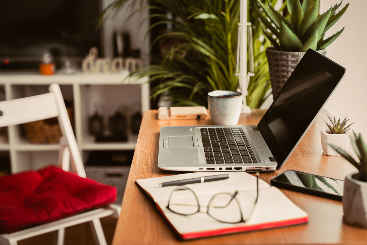 Close up view of a wooden desk with laptop, planner, mobile phone and eyeglasses left on the work table. Work from home concept with window light and green plants during quarantine and isolation