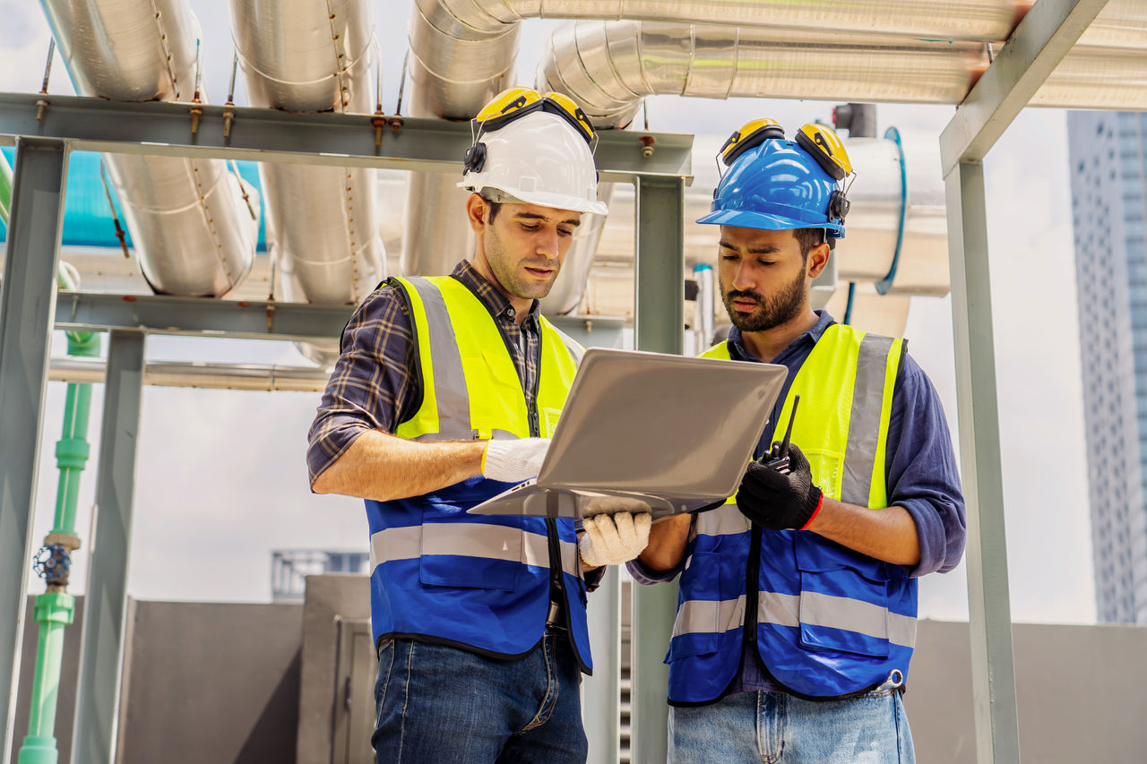 Collaboration, Two young construction workers discuss plans while reviewing blueprints on a rooftop. They wear safety gear and oversee intricate piping systems amidst a bustling urban backdrop,